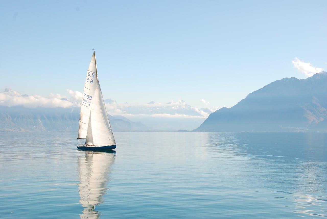 our-stories Sailing boat glides peacefully on a misty lake surrounded by mountains, under clear daylight.